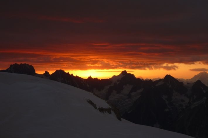 Ein dunkelroter / orangener Sonnenaufgang über mehreren Bergspitzen. Im Vordergrund eine weite Gletscherfläche.