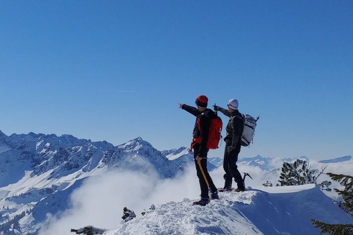 Zwei Alpinisten stehen auf einem schneebedeckten Grat und zeigen synchron mit dem rechten Zeigefinger in die gleiche Richtung.