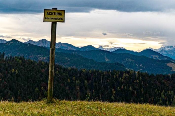 Gewittrige Bergumgebung mit Abbruchkante. An der Kante steht ein gelbes Schild mit der Aufschrift "ACHTUNG Absturzgefahr!"