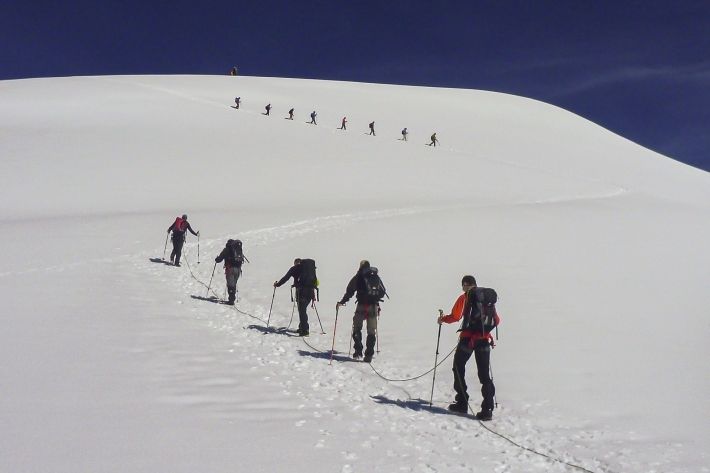 Zwei Seilschaften mit 5 und 8 Personen laufen unterhalb eines schneebedeckten Gipfel im Zickzack aufeinander zu.